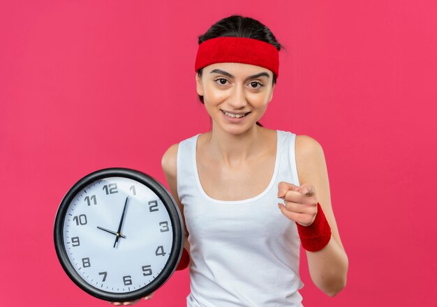 Junge Fitnessfrau in Sportbekleidung mit Stirnband, das Wanduhr hält, zeigt mit Index zur Kamera mit Lächeln auf Gesicht, das über rosa Wand steht