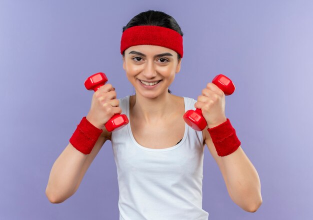 Junge Fitnessfrau in Sportbekleidung mit Stirnband, das zwei Hanteln in erhöhten Händen hält