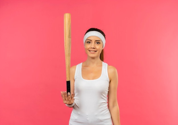 Junge Fitnessfrau in Sportbekleidung mit Stirnband, die eine Fledermaus mit Lächeln auf Gesicht hält, das über rosa Wand steht