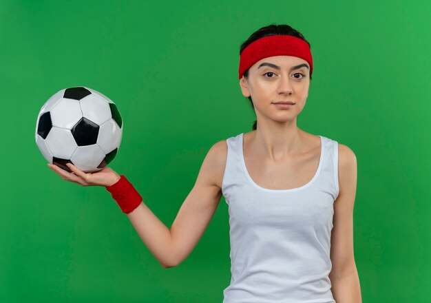 Junge Fitnessfrau in Sportbekleidung mit Stirnband, die Fußball mit dem sicheren Ausdruck steht, der über grüner Wand steht