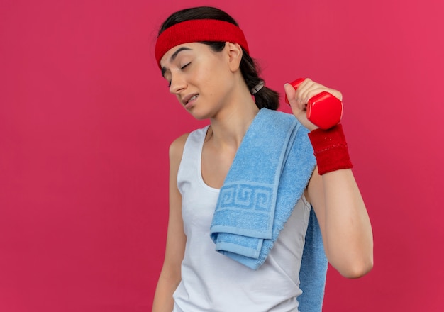 Junge Fitnessfrau in Sportbekleidung mit Stirnband, die Hantel in der erhöhten Hand hält, die Übungen macht, die müde und gelangweilt stehen über rosa Wand stehen