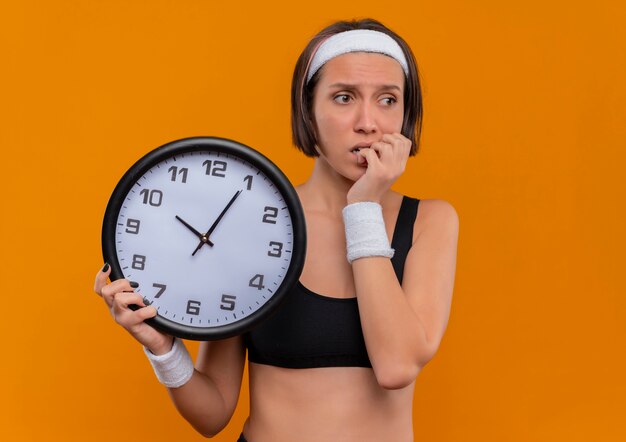 Junge Fitnessfrau in Sportbekleidung mit Stirnband, die Wanduhr hält, die beiseite gestresste und nervöse beißende Nägel steht, die über orange Wand stehen