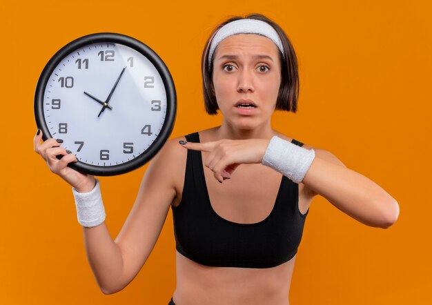 Junge Fitnessfrau in Sportbekleidung mit Stirnband, die Wanduhr hält, die mit Zeigefinger darauf zeigt, verwirrt und sehr besorgt stehend über orange Wand