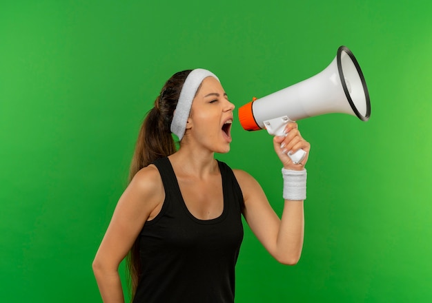 Kostenloses Foto junge fitnessfrau in sportbekleidung mit stirnband, die zu megaphon schreit, das über grüner wand steht