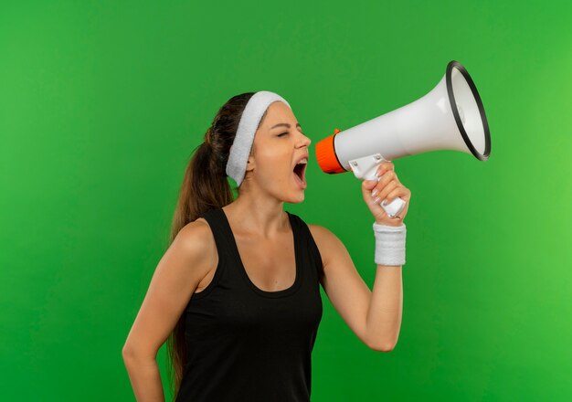 Junge Fitnessfrau in Sportbekleidung mit Stirnband, die zu Megaphon schreit, das über grüner Wand steht