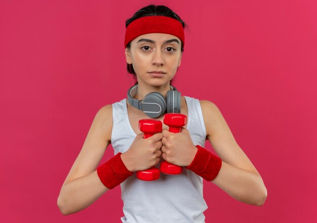 Junge Fitnessfrau in Sportbekleidung mit Stirnband, die zwei Hanteln hält, die Übungen mit ernstem Gesicht über rosa Wand stehen