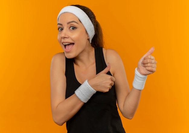 Junge Fitnessfrau in Sportbekleidung mit Stirnband glücklich und überrascht, mit den Fingern zur Seite stehend über orange Wand stehend