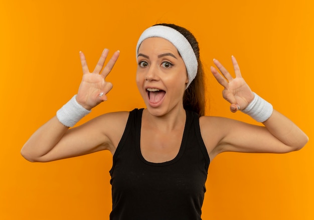 Junge Fitnessfrau in Sportbekleidung mit Stirnband lächelnd glücklich und positiv zeigt ok Zeichen, das über orange Wand steht