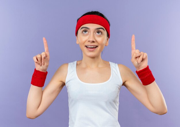 Junge Fitnessfrau in Sportbekleidung mit Stirnband lächelnd zuversichtlich mit Zeigefingern oben stehend über lila Wand