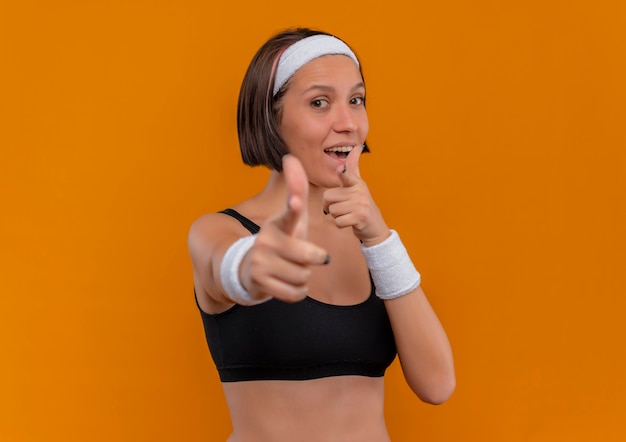 Junge Fitnessfrau in Sportbekleidung mit Stirnband positiv und glücklich mit Zeigefingern auf Kamera stehend über orange Wand