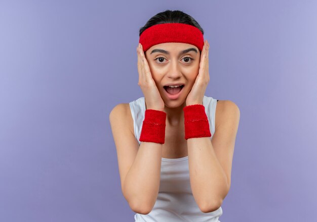 Junge Fitnessfrau in Sportbekleidung mit Stirnband schockiert mit Armen auf ihrem Gesicht, das über lila Wand steht