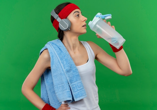 Junge fitnessfrau in sportbekleidung mit stirnband und handtuch auf ihrer schulter, die flasche wasser hält, das nach dem training steht, das über grüner wand steht