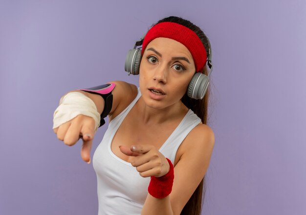 Junge Fitnessfrau in Sportbekleidung mit Stirnband und Kopfhörern, die mit dem Finger auf Kamera zeigen, besorgt über graue Wand stehend