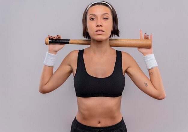 Junge Fitnessfrau in Sportkleidung, die Baseballschläger hält, der zuversichtlich weiß über weißer Wand steht