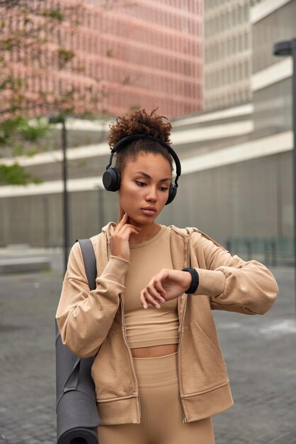 Junge Fitnessfrau mit lockigen Haaren überprüft den Puls auf der Smartwatch, nachdem das in Sportkleidung gekleidete Cardio-Training Karemat trägt, hört Musik über Kopfhörer, posiert im Freien vor verschwommenem Hintergrund