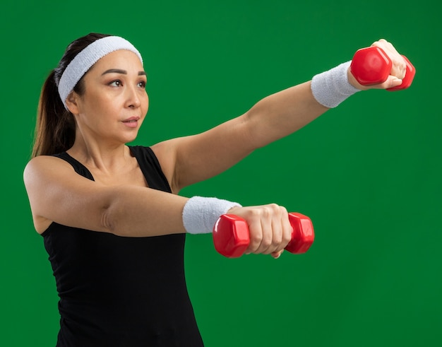 Junge Fitnessfrau mit Stirnband mit Hanteln, die angespannte und selbstbewusste Übungen über grüner Wand macht