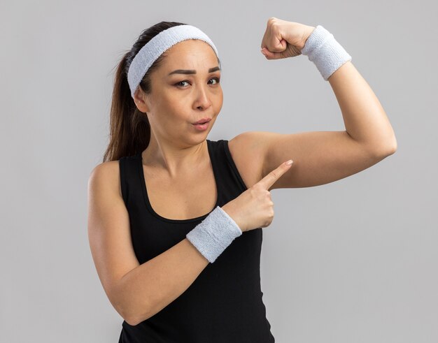 Junge Fitnessfrau mit Stirnband und Armbinden, die die Faust heben und Stärke und Kraft zeigen, die über weißer Wand steht