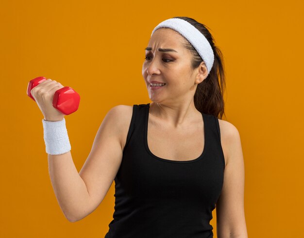 Junge Fitnessfrau mit Stirnband und Armbinden, die eine Hantel hält und sie verwirrt und unzufrieden über orangefarbener Wand steht