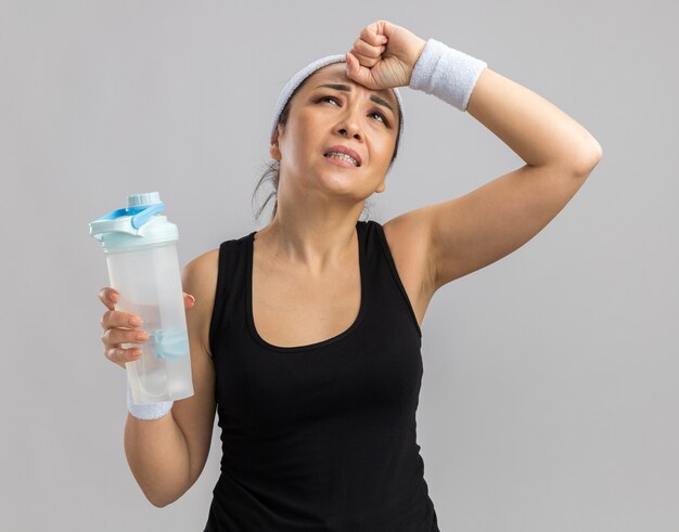 Junge Fitnessfrau mit Stirnband und Armbinden, die eine Wasserflasche hält und mit der Hand auf dem Kopf verwirrt aussieht, weil sie über der weißen Wand steht