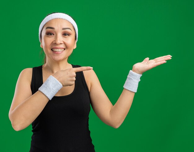 Junge Fitnessfrau mit Stirnband und Armbinden mit einem Lächeln im Gesicht, die mit dem Arm der Handkopie präsentiert, der mit dem Zeigefinger zur Seite zeigt