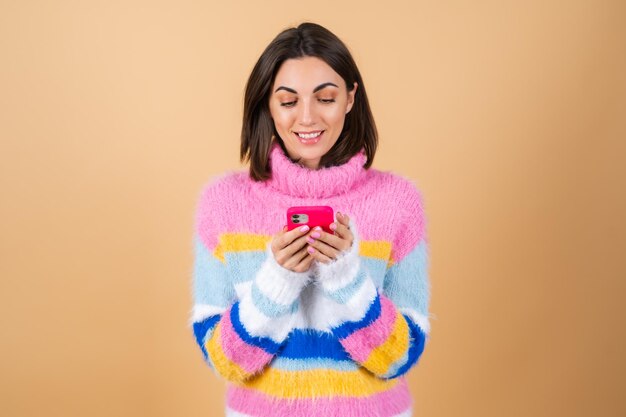 Junge Frau auf Beige in einem Strickpullover mit einem Telefon schaut auf den Bildschirm und lächelt