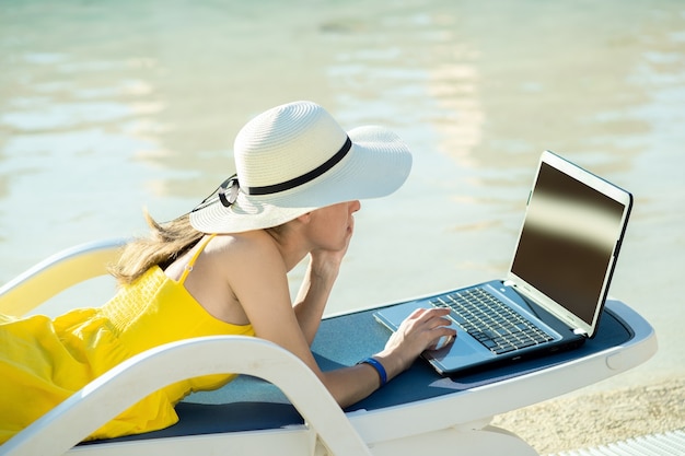Junge frau auf strandkorb am schwimmbad, das am computer-laptop arbeitet