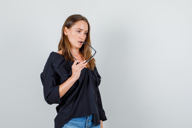 Junge Frau beißt Brille beim Denken im Hemd, kurze Vorderansicht.