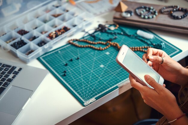 Junge Frau chattet mit ihrem Handy, während sie mit ihrem Schmuckhobby neben dem Tisch sitzt.