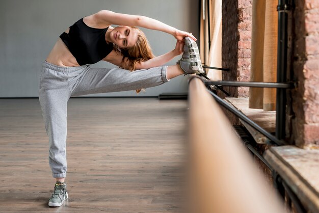 Junge Frau der Eignung, die sein Bein mithilfe von Barre im Tanzstudio ausdehnt