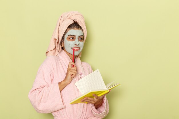 Junge Frau der Vorderansicht im rosa Bademantel, die Notizen schreibt, die auf einer grünen Oberfläche denken