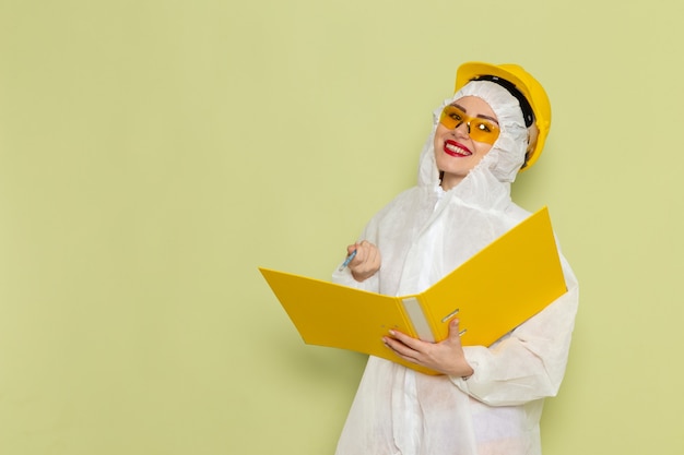 Junge Frau der Vorderansicht im weißen Sonderanzug und im gelben Helm, der gelbe Dateien hält, die auf der einheitlichen Wissenschaft des Grünraumanzugs lächeln