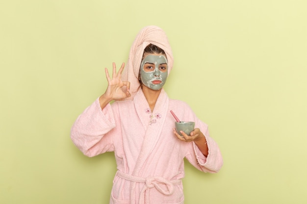 Junge Frau der Vorderansicht nach der Dusche im rosa Bademantel, der Maske auf einem grünen Schreibtisch anwendet