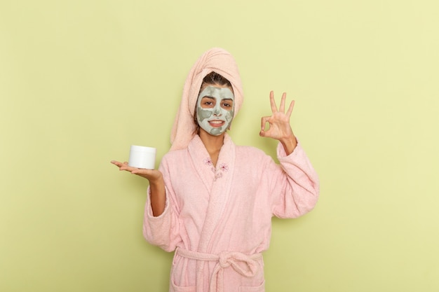 Junge Frau der Vorderansicht nach Dusche im rosa Bademantel, der Creme hält und auf einer grünen Oberfläche lächelt