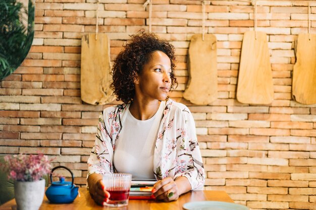 Junge Frau des Afroamerikaners, die im Restaurant mit Cocktail auf Holztisch sitzt