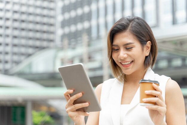 Junge Frau des erfolgreichen schönen asiatischen Geschäfts, die in der Hand intelligentes Telefon und Kaffeetasse verwendet