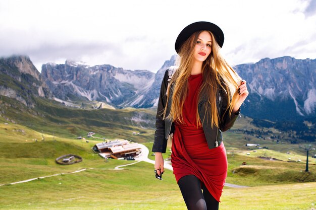 Junge Frau, die am Alpengebirge aufwirft, Kleid, Lederjacke, Sonnenbrille und Rucksack tragend