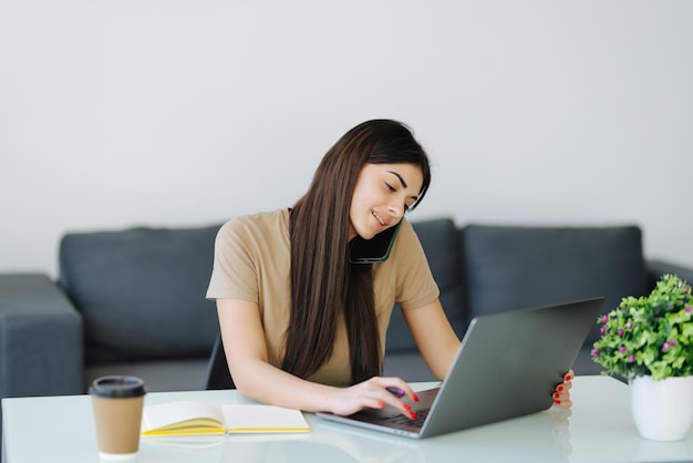 Junge Frau, die am Laptop arbeitet, lächelt, spricht am Telefon, Sonne kommt durchs Fenster