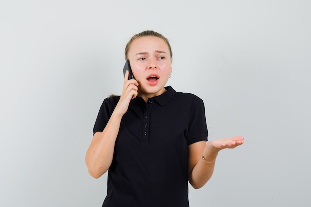 Junge Frau, die am Telefon spricht und ihre eine Hand im schwarzen T-Shirt hebt und frustriert aussieht