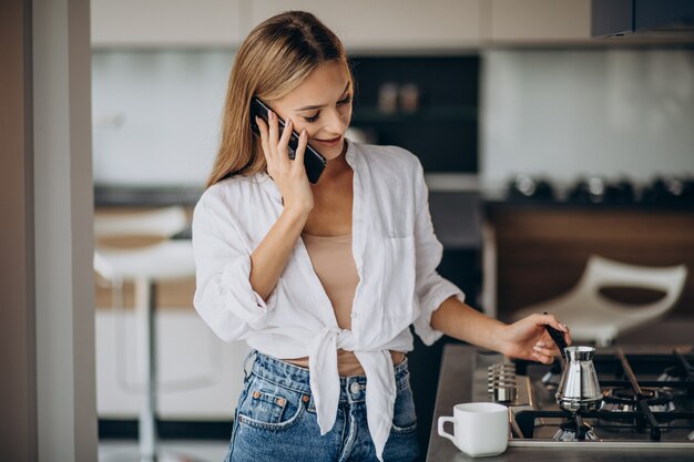 Junge Frau, die am Telefon spricht und Morgenkaffee kocht