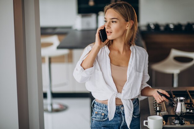 Junge Frau, die am Telefon spricht und Morgenkaffee kocht