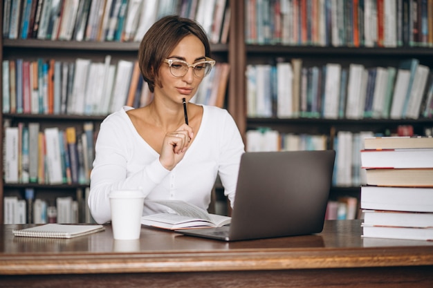 Junge Frau, die an der Bibliothek unter Verwendung der Bücher und des Computers sitzt