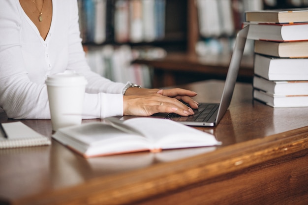 Junge Frau, die an der Bibliothek unter Verwendung der Bücher und des Computers sitzt