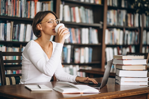 Junge Frau, die an der Bibliothek unter Verwendung der Bücher und des Computers sitzt