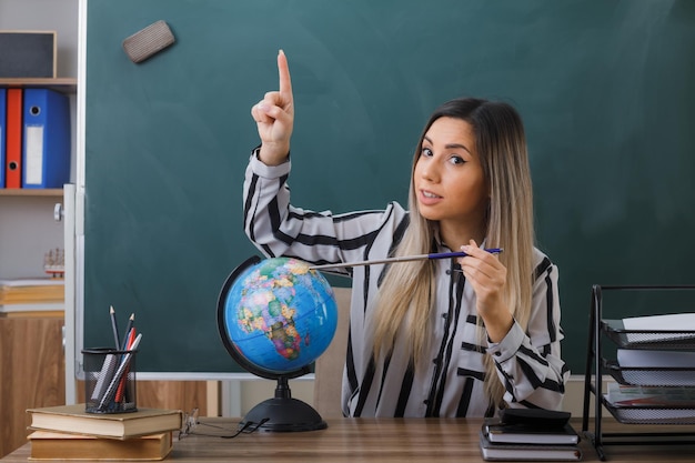 junge frau, die an der schulbank vor der tafel im klassenzimmer sitzt und den unterricht erklärt, der globus und zeiger hält und selbstbewusst lächelnd aussieht