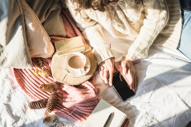 Junge Frau, die auf Bett mit Tasse Kaffee liegt und Smartphone verwendet