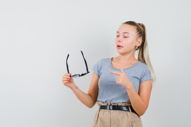 Junge Frau, die auf Brille im T-Shirt, in der Hose zeigt und zuversichtlich schaut. Vorderansicht.