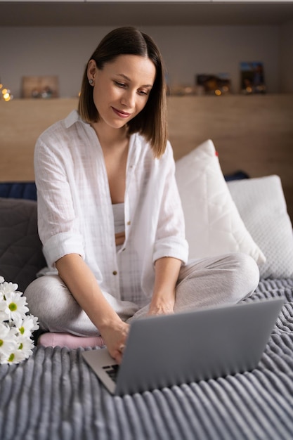 Junge Frau, die auf dem Bett sitzt und Pyjamas trägt, genießt weiße Blumen und plaudert mit dem Laptop