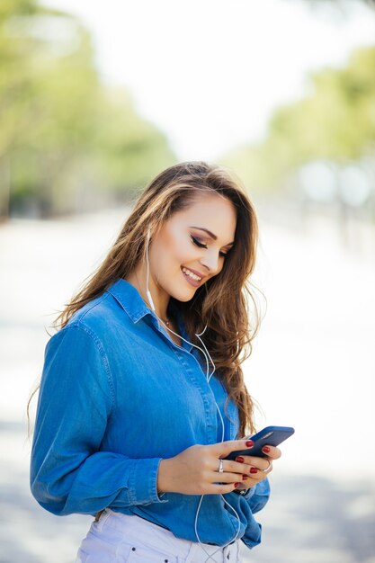 Junge Frau, die auf dem Smartphone, das in der Straße an einem sonnigen Tag geht, SMS schreibt