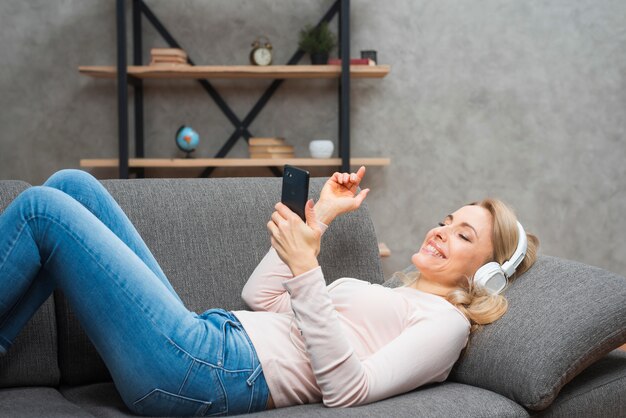 Junge Frau, die auf dem Sofa genießend liegt, die Musik auf Kopfhörer von einem intelligenten Telefon hörend