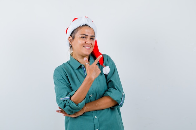 Junge Frau, die auf die obere rechte Ecke im Hemd, in der Weihnachtsmütze zeigt und fröhlich schaut. Vorderansicht.